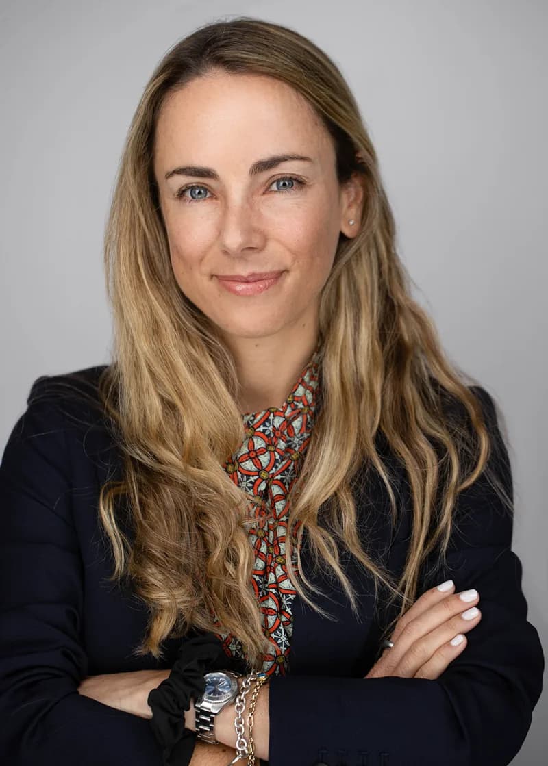 Professional headshot of woman with long blonde hair wearing a navy blazer with a patterned blouse against a gray background