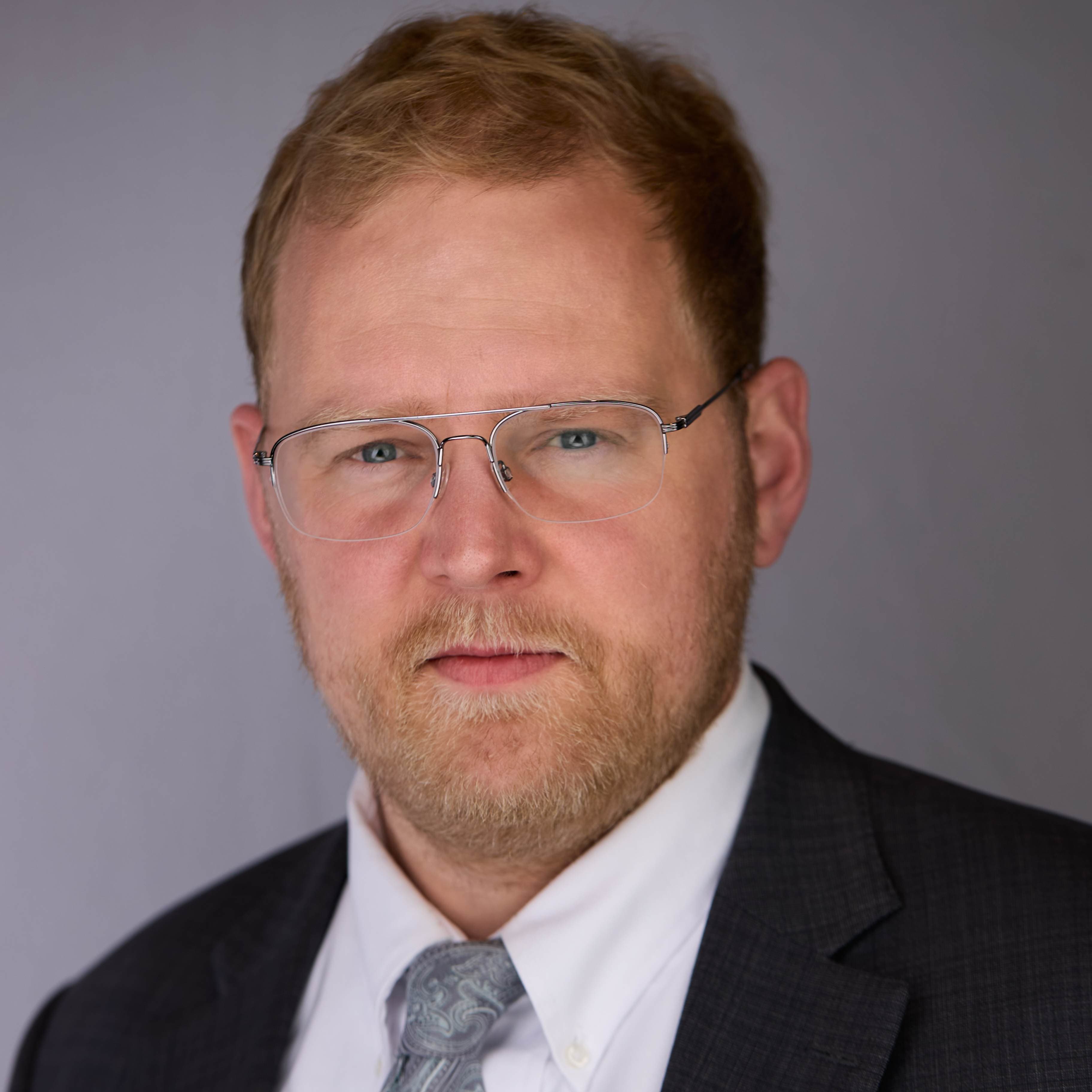 Professional headshot of Cameron Hansen in a dark suit with glasses and a patterned tie