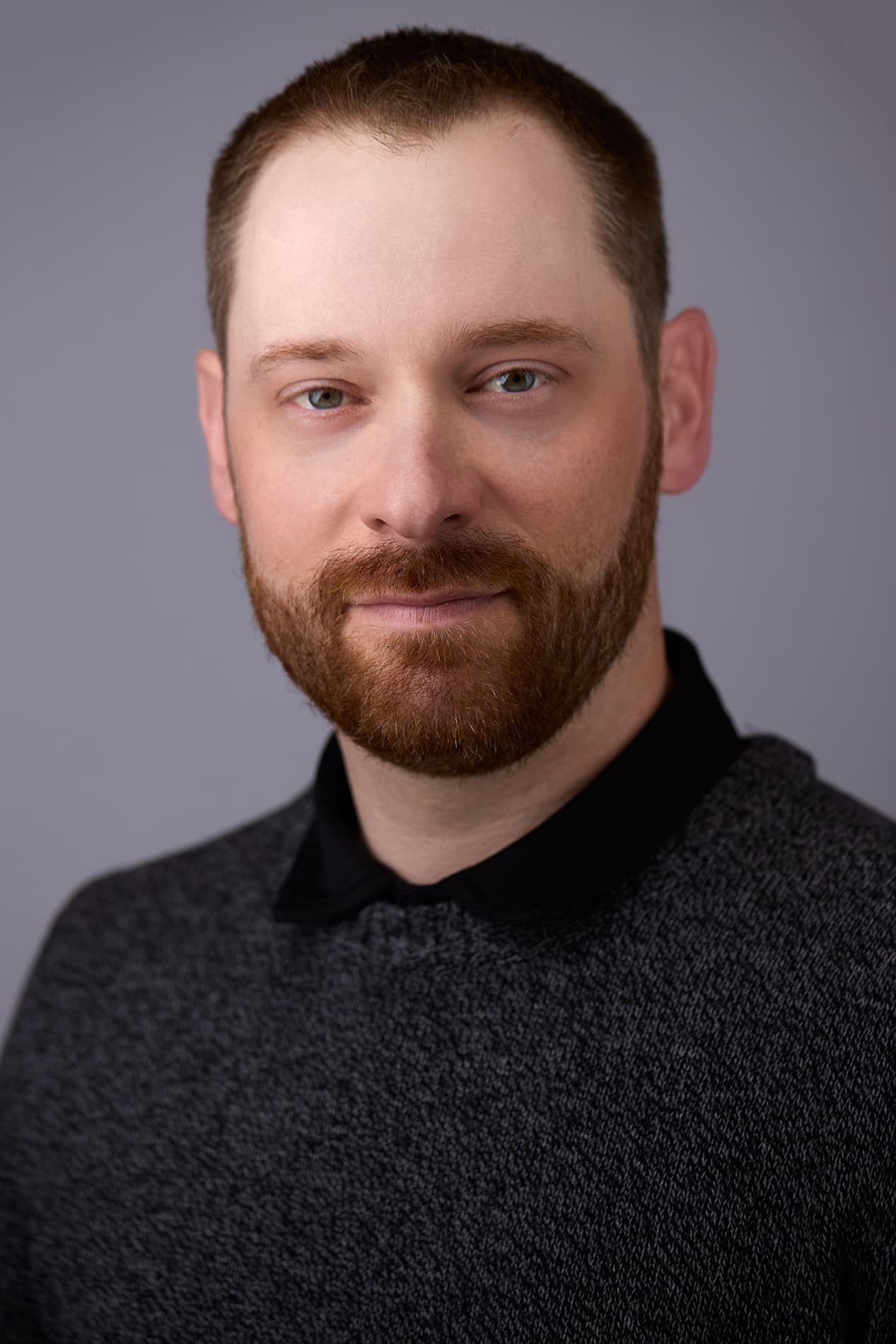 Professional headshot of man with reddish-brown hair and beard in dark collared shirt against gray background