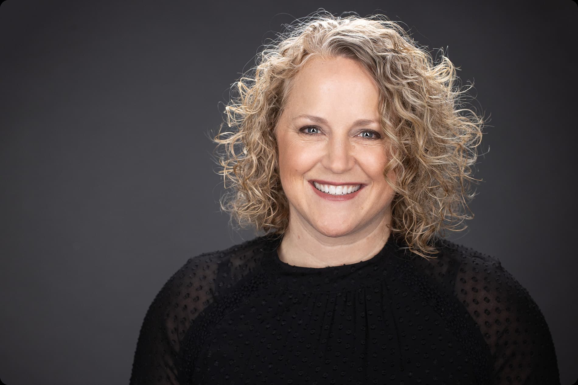 Professional headshot of woman with curly blonde hair in black textured top against dark background