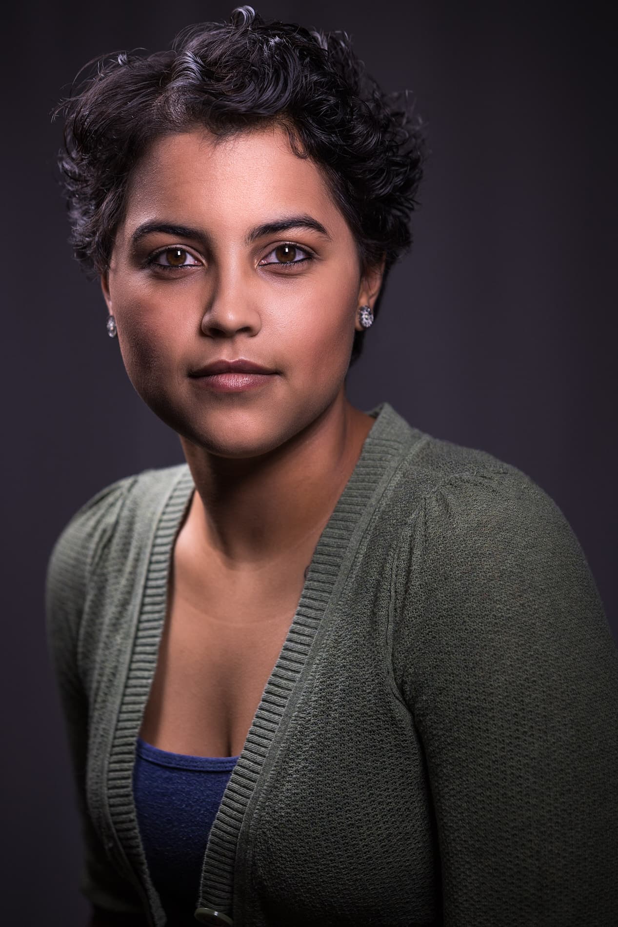 Professional headshot of woman with short curly dark hair in green cardigan over blue top against dark background