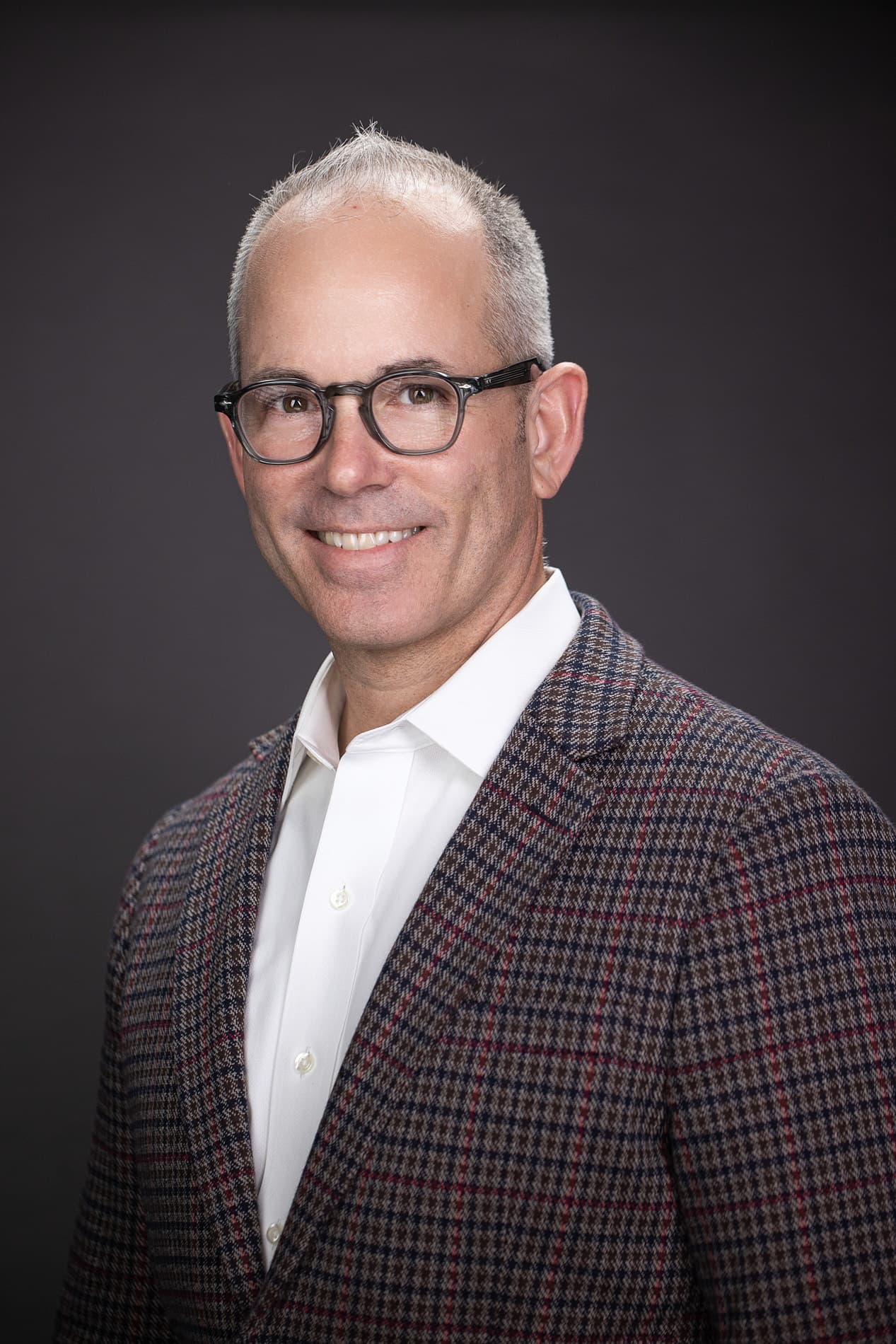 Professional headshot of an older man with gray hair, glasses, wearing a patterned suit jacket and white shirt against a dark background