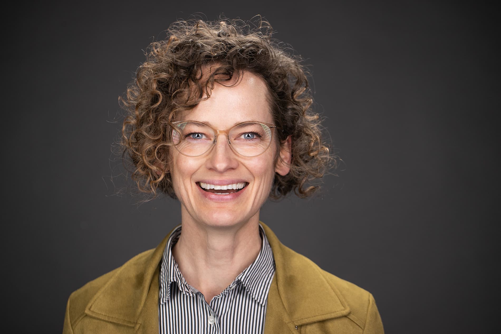Professional headshot of a woman with short curly hair and glasses wearing a striped shirt and mustard-colored jacket against a dark gray background