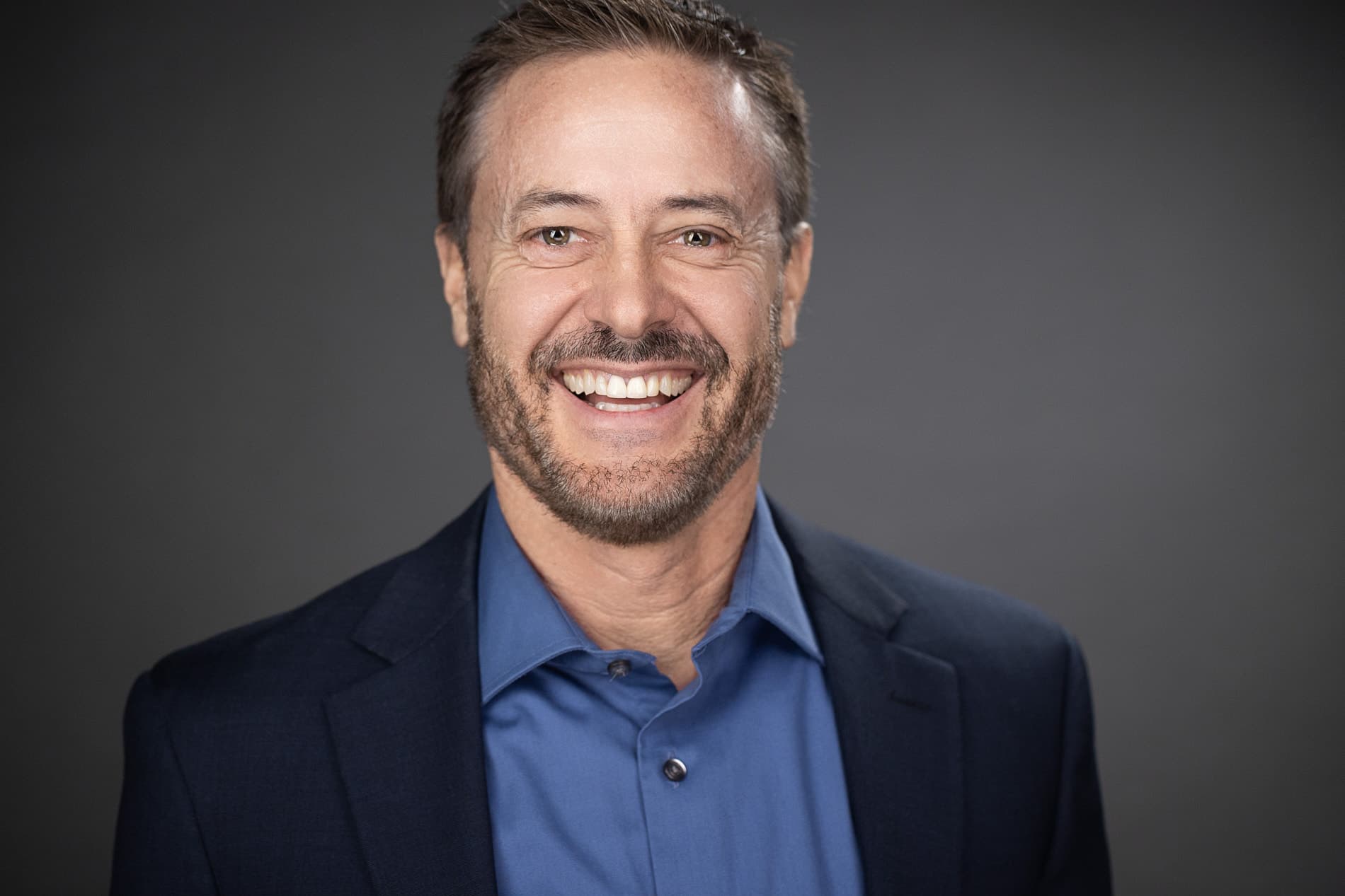 Professional headshot of a smiling man with beard and short hair in a navy suit and blue shirt against a dark gray background