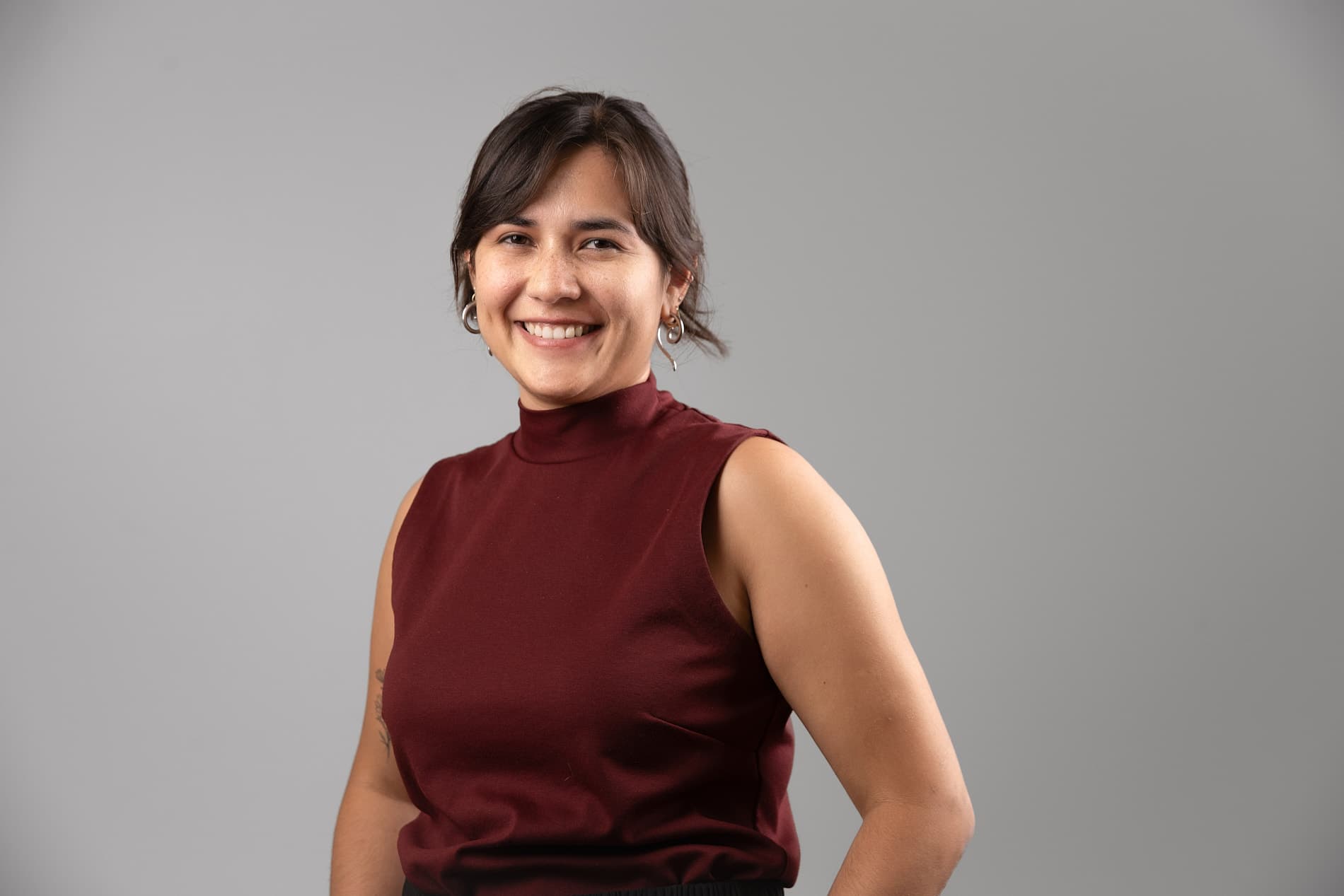 Professional headshot of young woman with dark hair in a burgundy sleeveless turtleneck top against a gray background