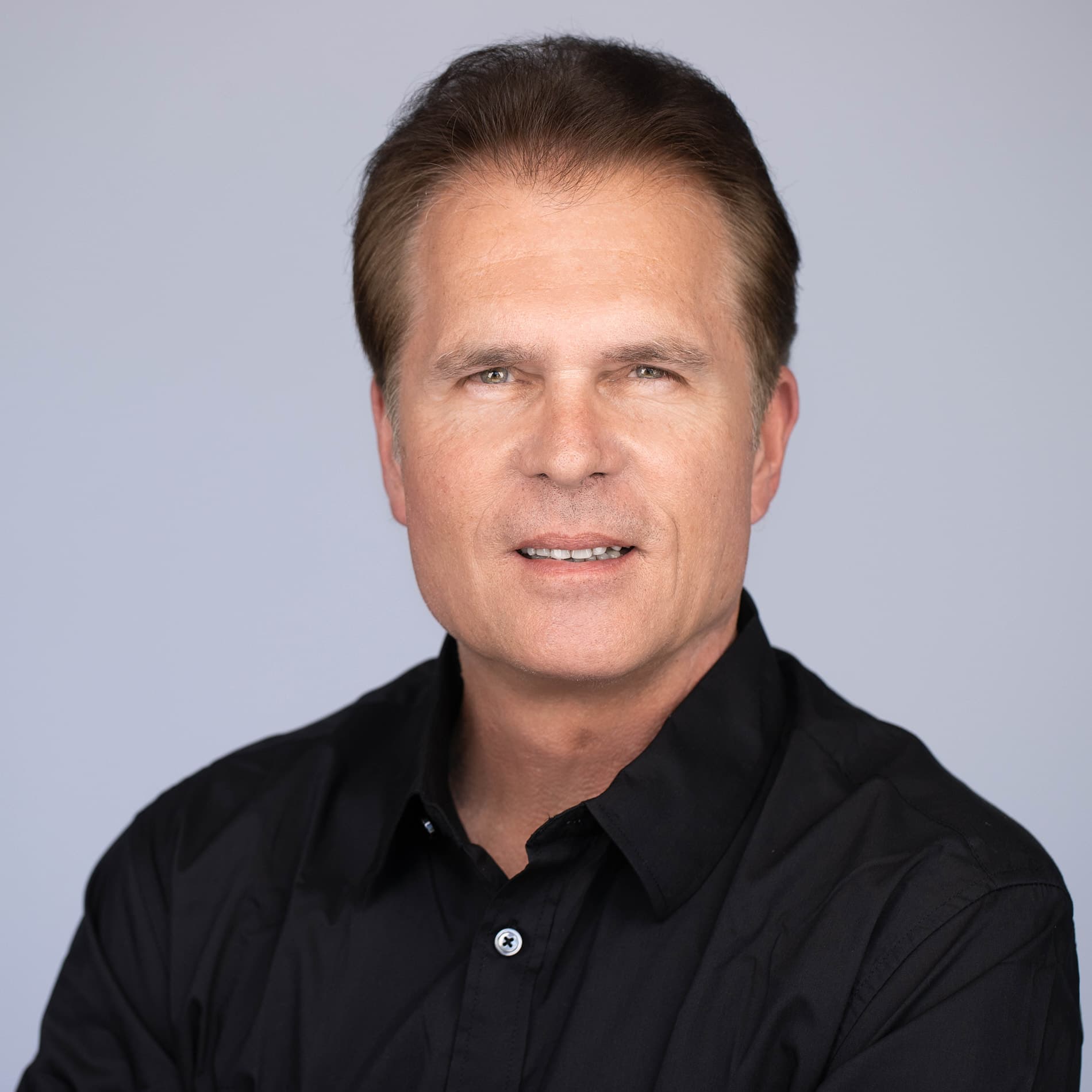 Professional headshot of man with brown hair in a black button-up shirt against a light blue background