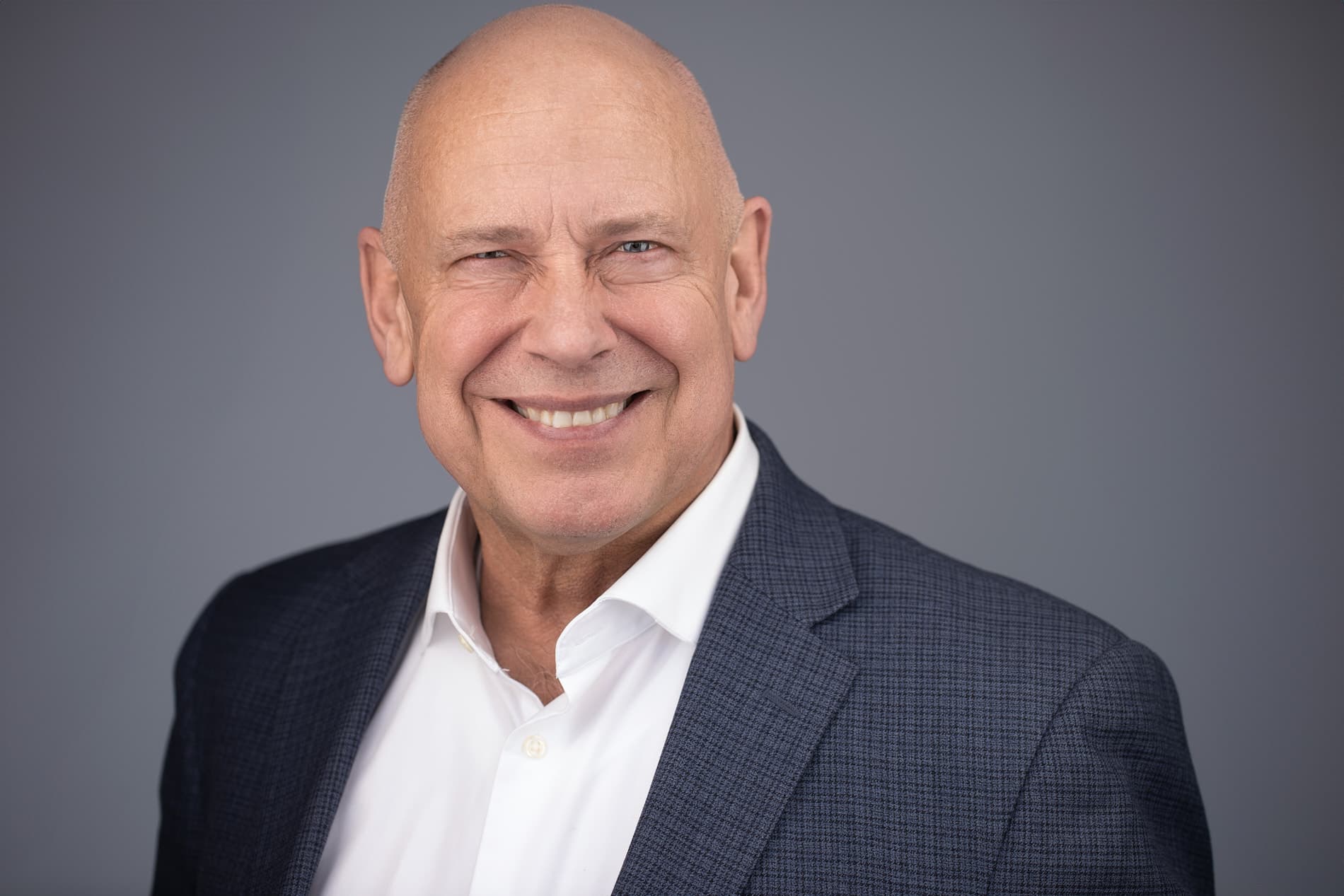 Professional headshot of bald man with a big smile wearing a navy patterned suit jacket and white shirt against a gray background