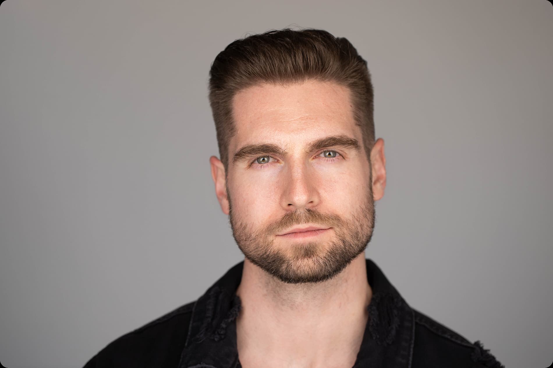 Professional headshot of man with styled brown hair and beard in black shirt against gray background