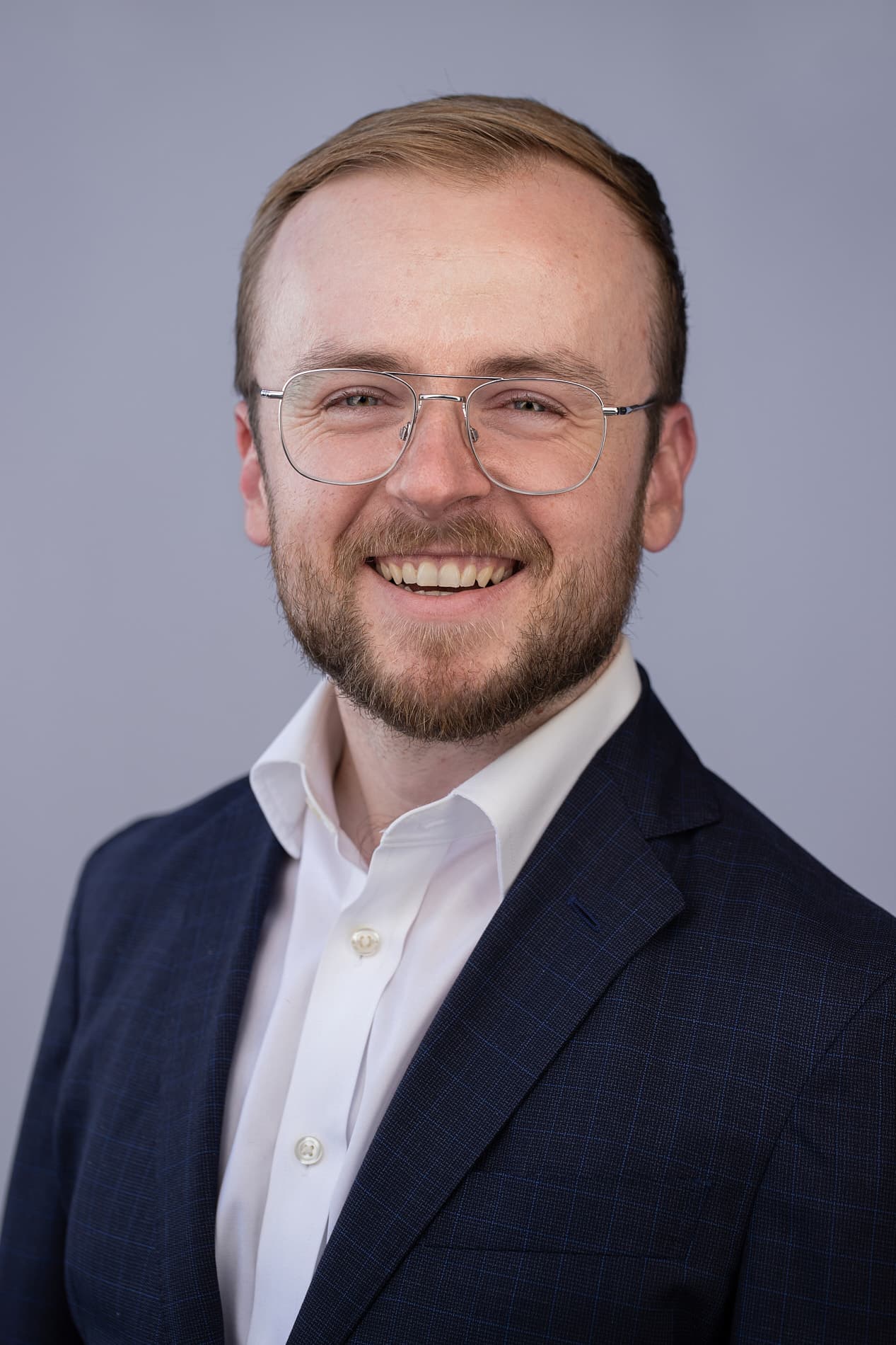 Professional headshot of man with light brown hair, beard and glasses in navy suit and white shirt against gray background