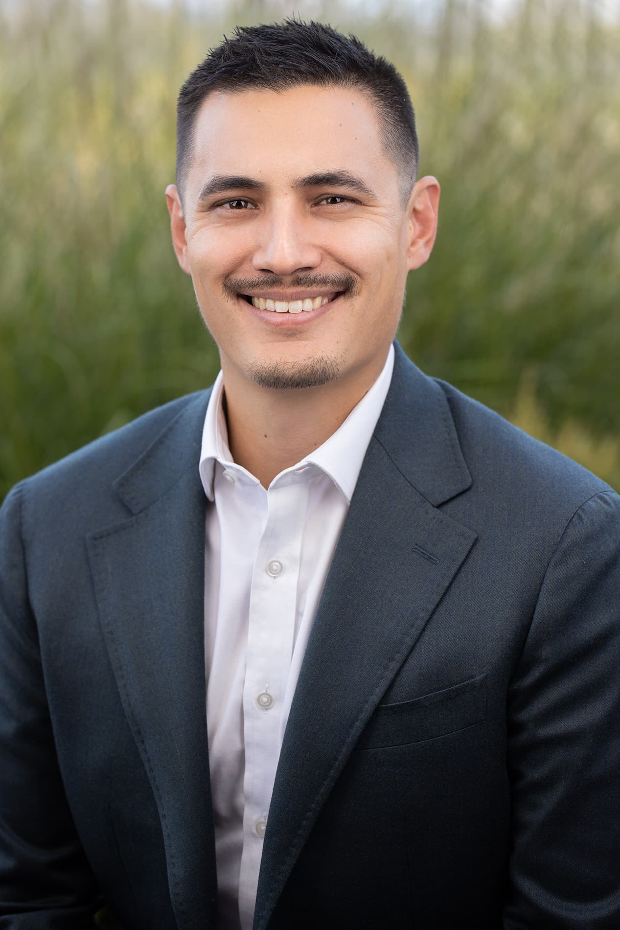 Professional headshot of young man in dark suit and white shirt against outdoor greenery background