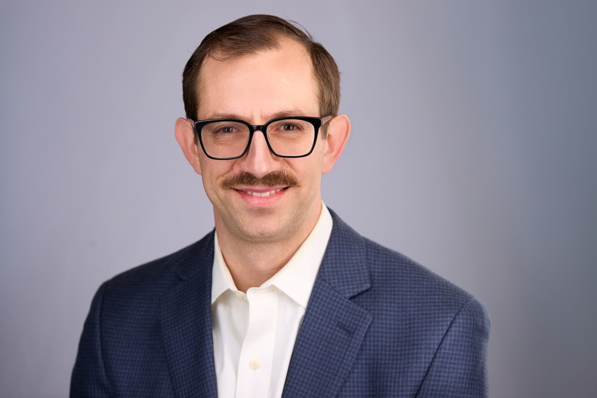 Professional headshot of a man with glasses and mustache in a navy patterned suit jacket and white shirt against a light gray background