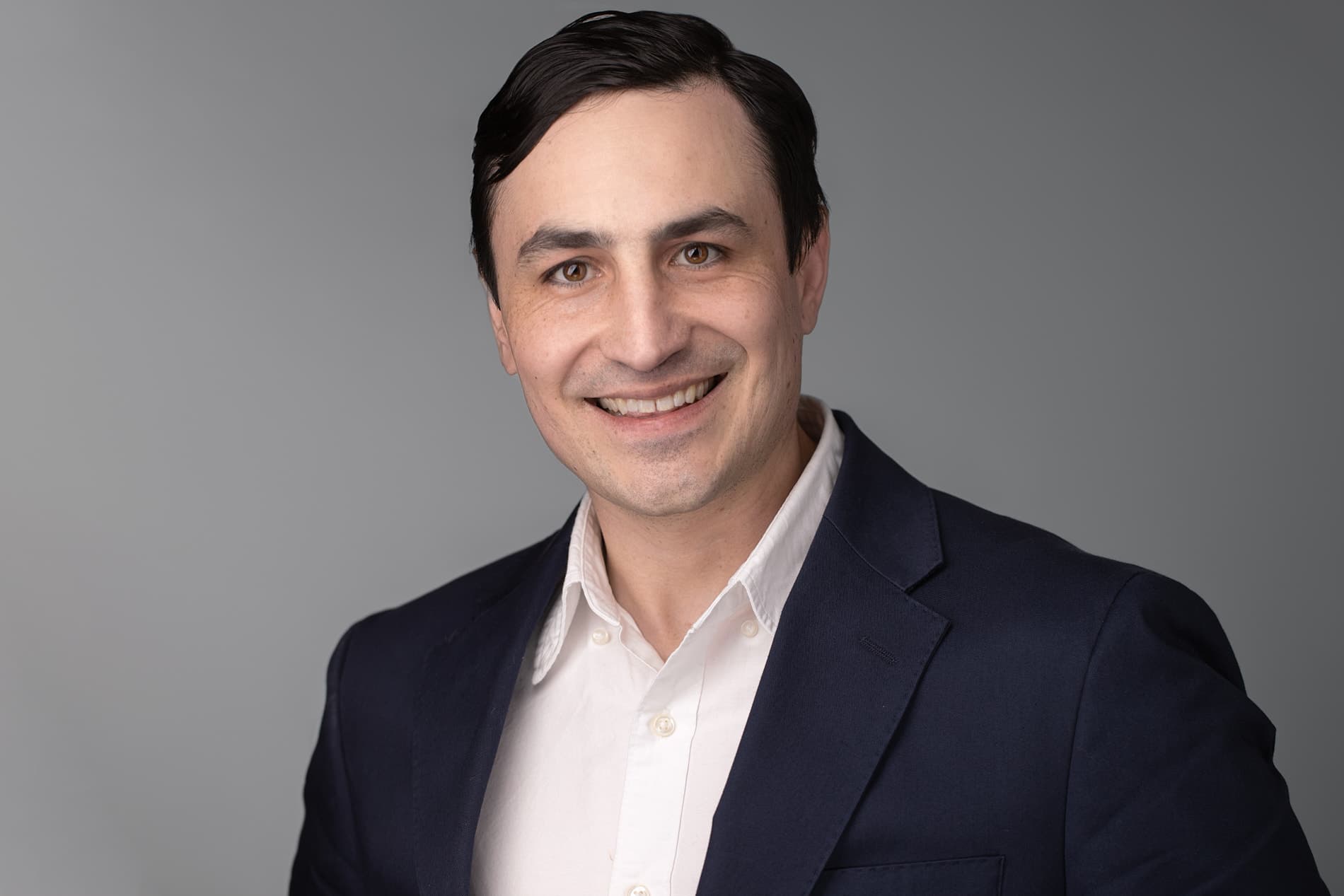 Professional headshot of a smiling man with dark hair in a navy suit and white shirt against a gray background