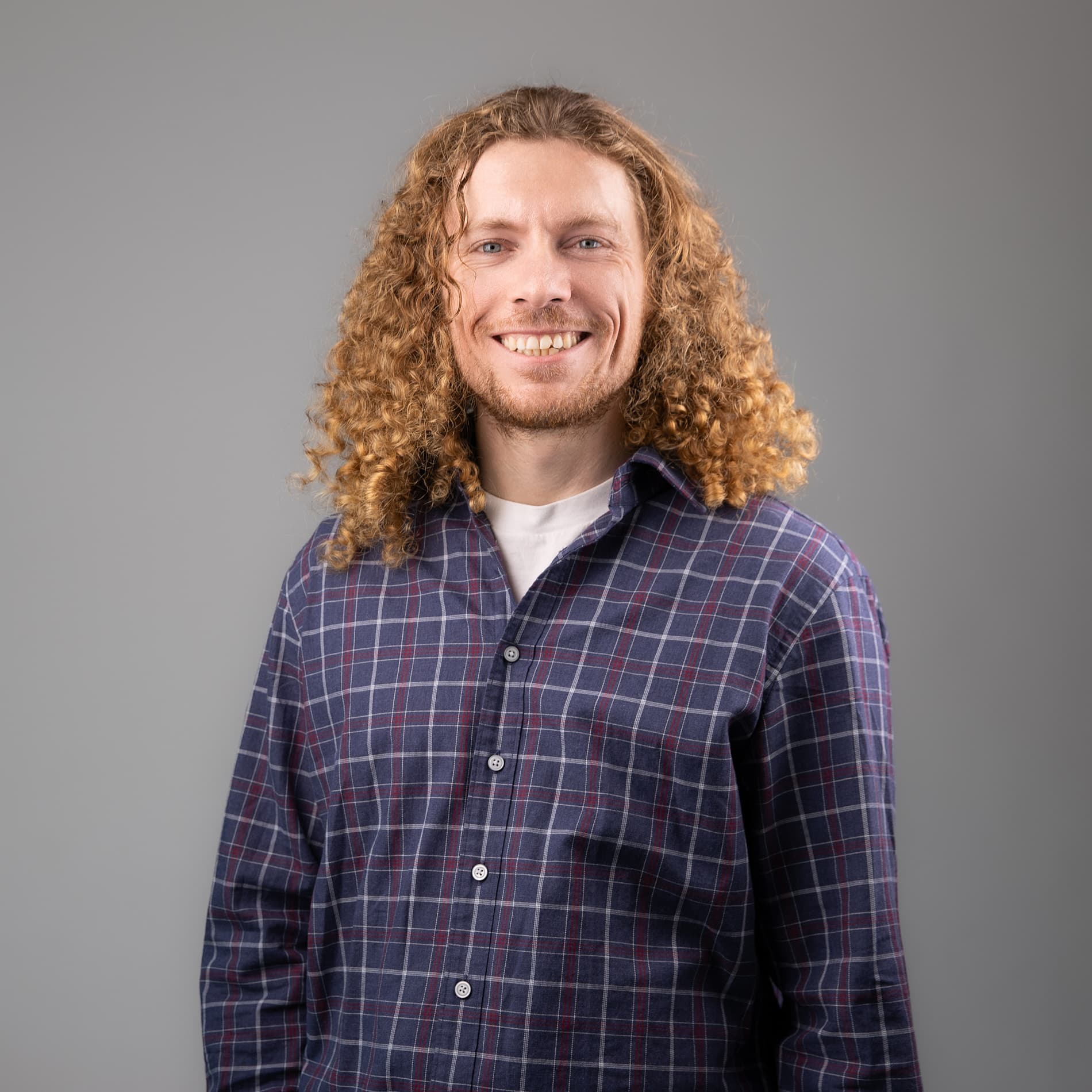 Professional headshot of a man with long curly blonde hair in a plaid button-up shirt against a gray background
