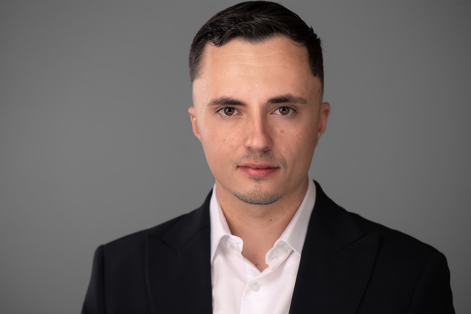 Professional headshot of a young man with short dark hair in a black suit and white shirt against a gray background