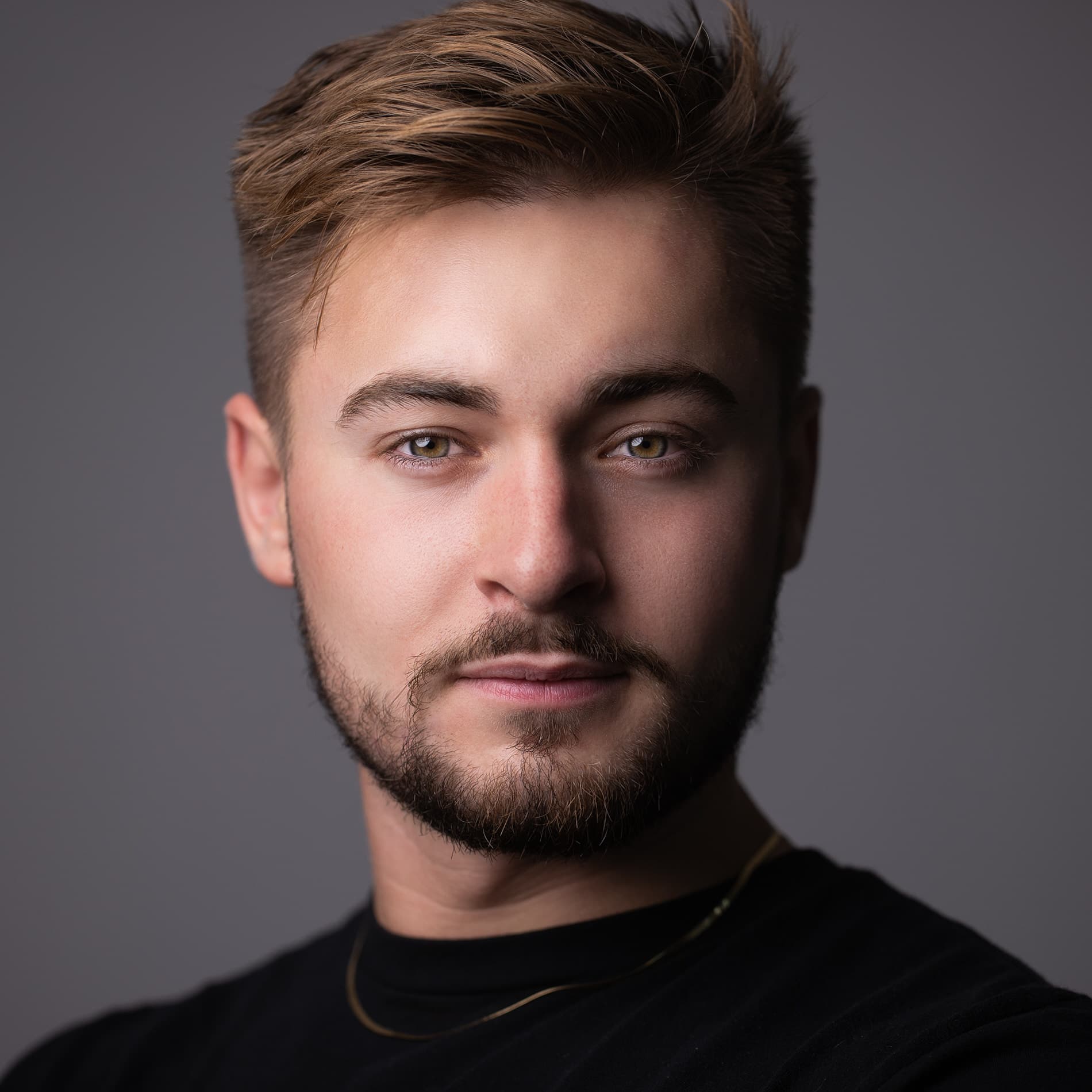 Professional headshot of a young man with styled brown hair and beard in a black shirt against a dark gray background
