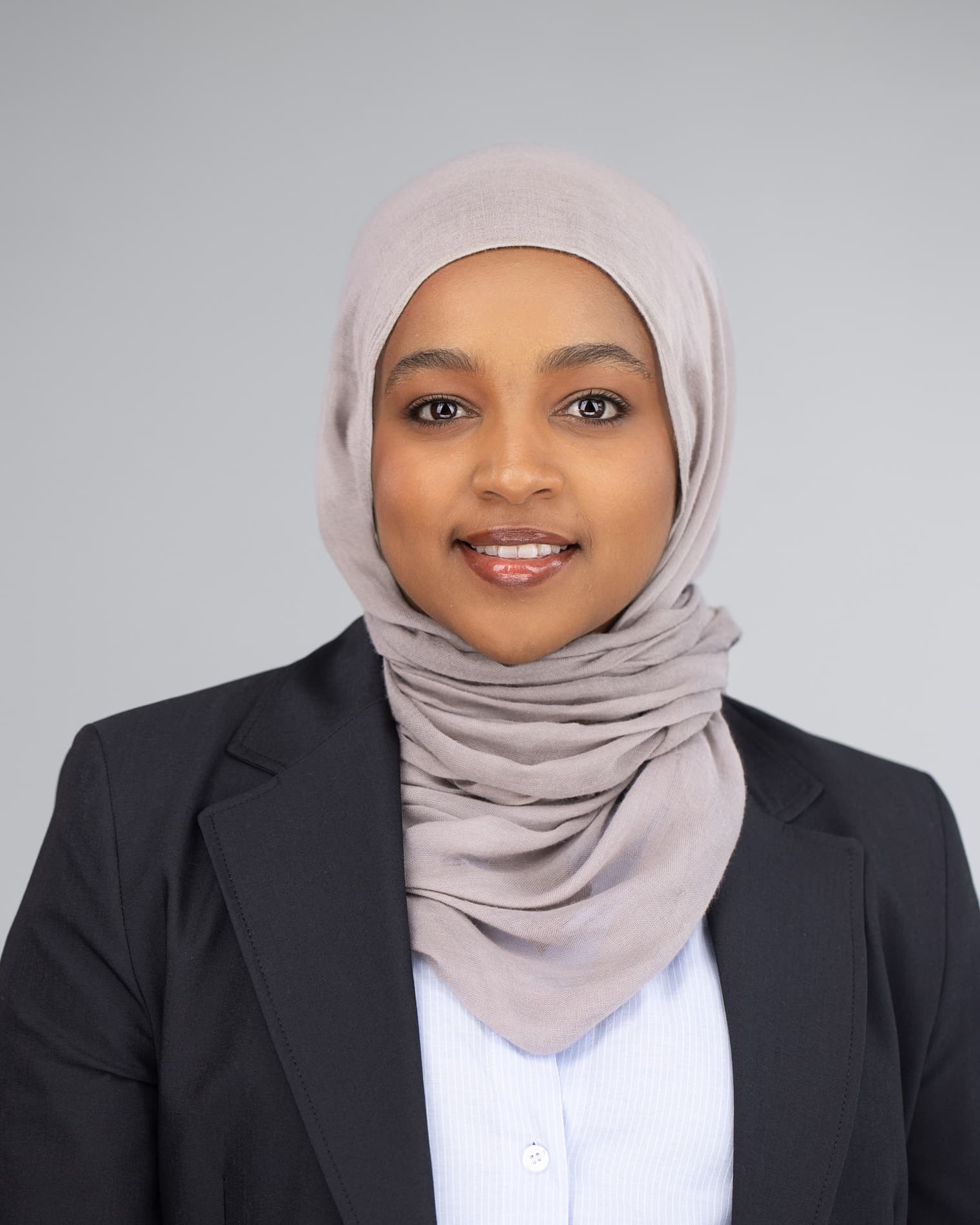 Professional headshot of young woman wearing a light gray hijab, white shirt and black blazer against a light gray background