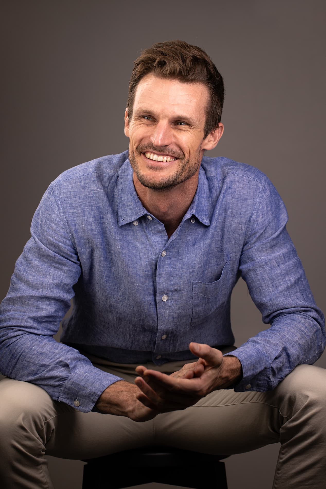 Professional headshot of man with brown hair and beard in a blue linen shirt against a gray background