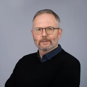 Professional headshot of a middle-aged man with glasses and a beard wearing a black sweater over a navy shirt