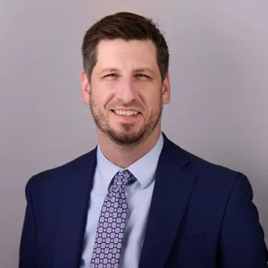 Professional headshot of a man with short brown hair and a beard wearing a navy blue suit with a patterned tie