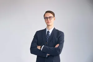 Professional headshot of a young man with glasses wearing a navy blue suit with a patterned tie