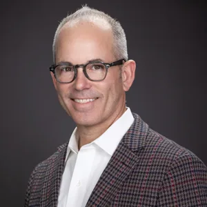 Professional headshot of a middle-aged man with gray hair, glasses wearing a patterned blazer and white shirt
