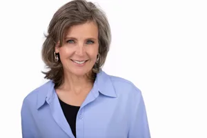 Professional headshot of a middle-aged woman with shoulder-length gray/brown hair wearing a light blue button-up shirt